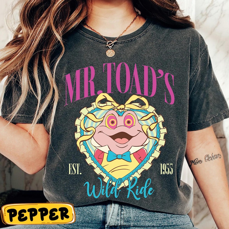 May include: Dark gray t-shirt featuring a cartoon frog character inside a heart-shaped design. The text "MR. TOAD'S" arches above the frog, with "Wild Ride" below. The shirt also includes the year "1955".
