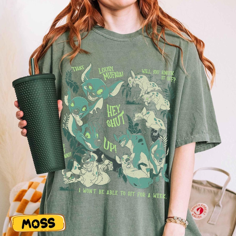 May include: Olive green t-shirt featuring a cartoon graphic of hyenas with text that reads "That Lousy Mufasa! Hey Shut Up! Will You Knock It Off?" and "I won't be able to sit for a week." A dark green studded tumbler is held in the image.