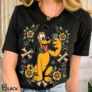 Op de afbeelding: Zwart t-shirt met een gele cartoonhond die een selfie maakt. Het ontwerp bevat gele bloemen, pootafdrukken en botten. Het woord "BLACK" staat linksonder.