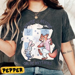 Pode incluir: T-shirt cinzento escuro com uma ilustra&ccedil;&atilde;o de desenho animado de personagens de Zootopia. O design inclui o texto "WHERE ANYONE CAN BE ANYTHING" e pegadas. A camisa tem um visual vintage e lavado.