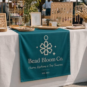 May include: A teal table runner featuring the white logo of Bead Bloom Co. with a flower icon, the text Charms, Keychains & Tiny Treasures, and EST. 2023. The table displays various keychains on metal stands and wooden boards with small trays.