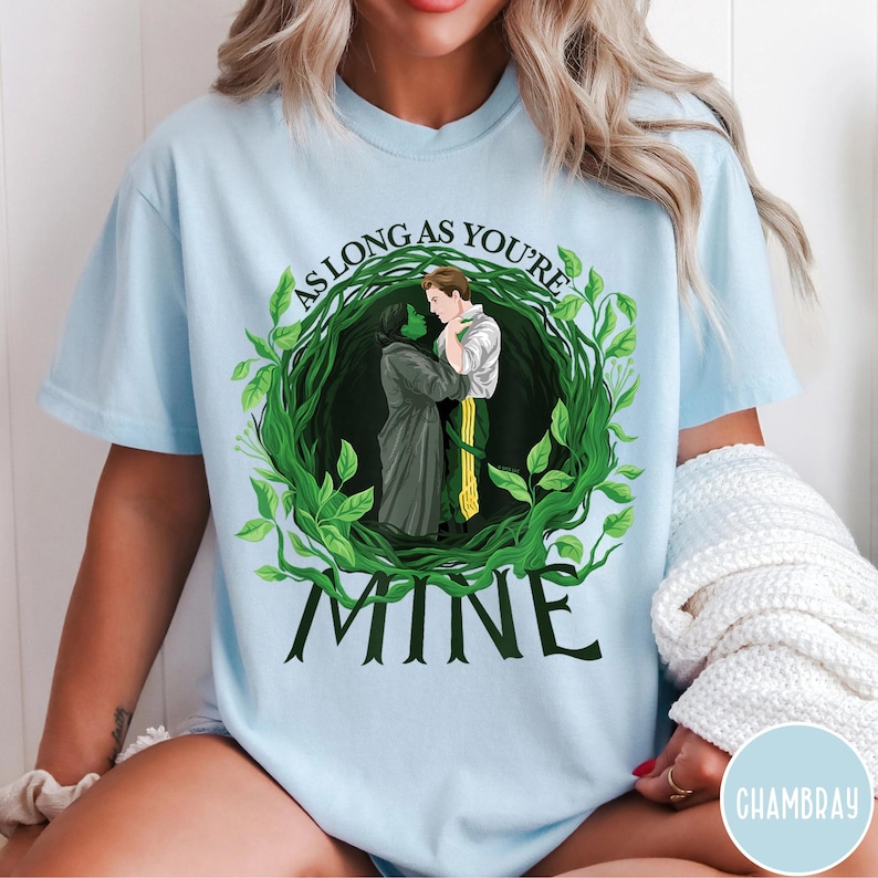 May include: Pale blue t-shirt with a graphic of two figures embracing, framed by a green leafy wreath. The text "AS LONG AS YOU'RE" curves above the image, with "MINE" below. The design is centrally placed on the shirt.