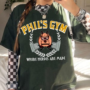 May include: Dark green t-shirt with the text "PHIL'S GYM" in yellow and white. The shirt features a cartoon character lifting weights, surrounded by a laurel wreath, and the text "WHERE HEROES ARE MADE".