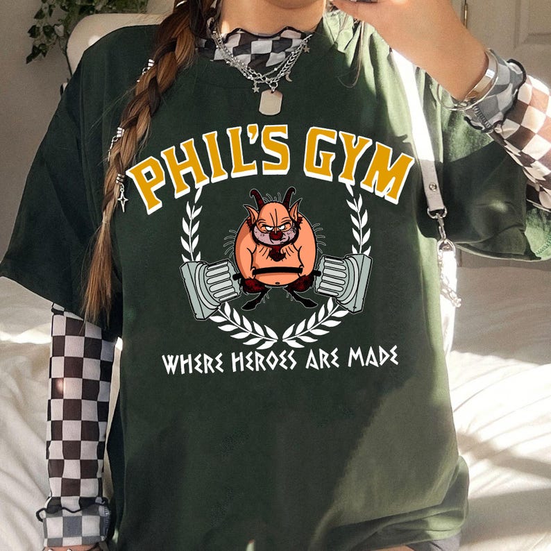 May include: Dark green t-shirt with the text "PHIL'S GYM" in yellow and white. The shirt features a cartoon character lifting weights, surrounded by a laurel wreath, and the text "WHERE HEROES ARE MADE".