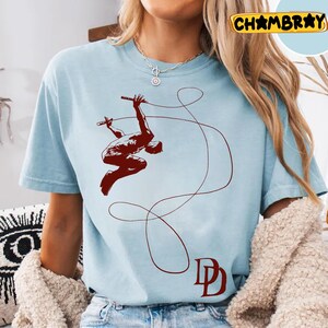 May include: Light blue t-shirt featuring a maroon graphic of a figure swinging on a rope, holding batons. The design includes a looping rope and the letters "DD" in maroon. The shirt is a casual wear item.