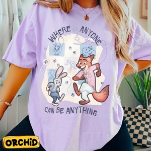 Pode incluir: Uma camiseta lil&aacute;s com personagens de Zootopia. A camiseta tem o texto "WHERE ANYONE CAN BE ANYTHING" com ilustra&ccedil;&otilde;es de Judy Hopps e Nick Wilde. A marca "ORCHID" est&aacute; impressa em um ret&acirc;ngulo amarelo na parte inferior.