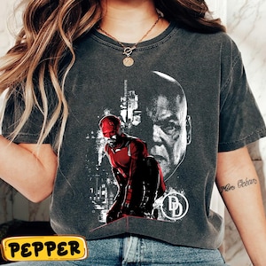 May include: Dark gray t-shirt featuring a graphic of a superhero in a red suit, kneeling in front of a cityscape. A large black and white portrait of a man is behind the superhero. The shirt also has a white logo.