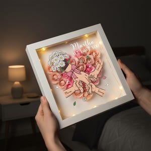 May include: A white framed artwork with a heart-shaped floral arrangement of pink roses and daisies. The artwork includes the text "Martha WE LOVE YOU" and illustrations of hands holding each other. Small fairy lights illuminate the interior.