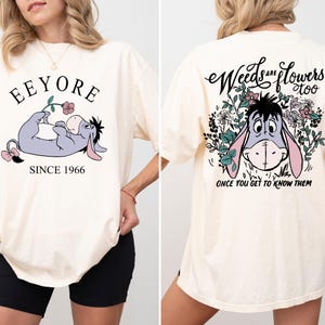 May include: A cream colored t-shirt with a cartoon image of Eeyore from Winnie the Pooh. The front of the shirt says "Eeyore Since 1966" and the back of the shirt says "Weeds are flowers too, once you get to know them".