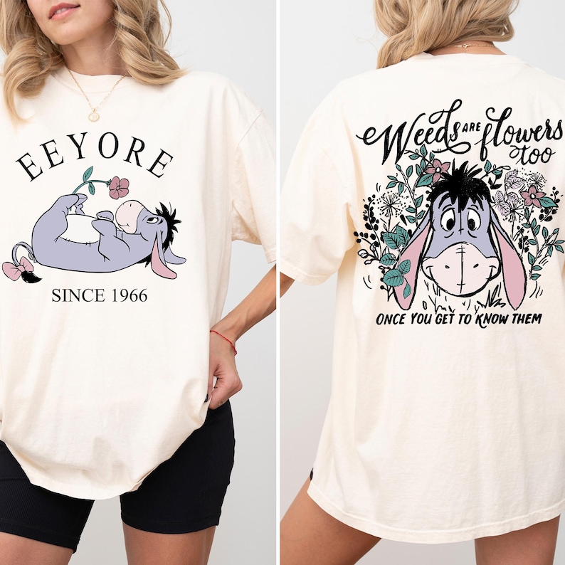 May include: A cream colored t-shirt with a cartoon image of Eeyore from Winnie the Pooh. The front of the shirt says "Eeyore Since 1966" and the back of the shirt says "Weeds are flowers too, once you get to know them".