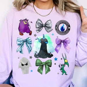 May include: A light purple sweatshirt featuring various cartoon character graphics and bow designs. The graphics include a purple monster, a blue flame, and a green creature. The bows are in silver, blue, green, and purple.