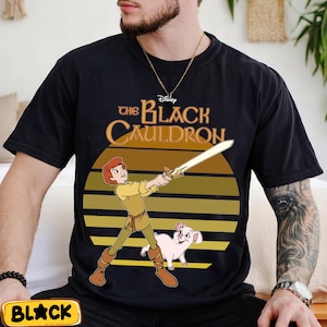 May include: Black t-shirt featuring a graphic from Disney's "The Black Cauldron." The design includes a cartoon illustration of a character holding a sword, a pig, and the film's title in gold lettering against a striped sunset background.