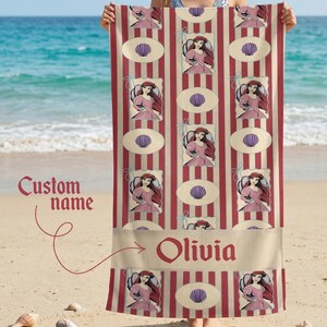 Op de afbeelding: Een strandlaken met een herhalend patroon van een tekenfilmprinses in een roze jurk, zeeschelpen en rood-cr&egrave;mekleurige strepen. De naam "Olivia" is in het rood onderaan gedrukt.