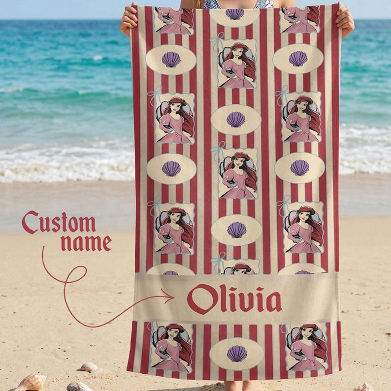 Op de afbeelding: Een strandlaken met een herhalend patroon van een tekenfilmprinses in een roze jurk, zeeschelpen en rood-cr&egrave;mekleurige strepen. De naam "Olivia" is in het rood onderaan gedrukt.