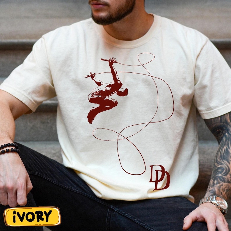 May include: Ivory-colored t-shirt featuring a maroon graphic of a figure swinging with batons and a rope. The design includes the letters "DD" in maroon. The shirt is short-sleeved.