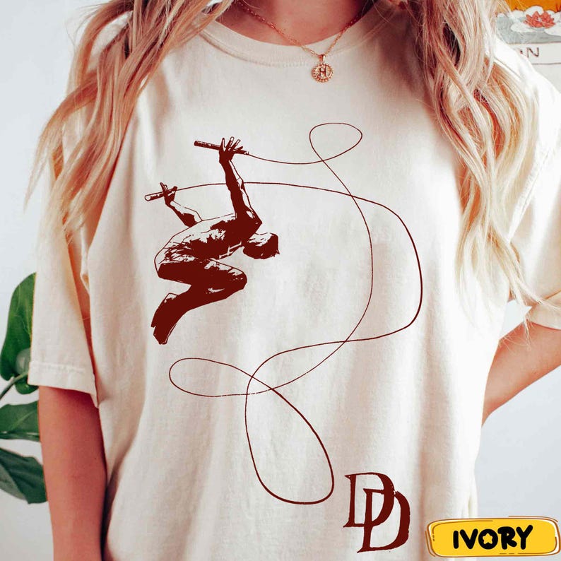 May include: Ivory-colored t-shirt featuring a brown illustration of a person swinging with a rope. The design includes the letters "DD" in brown at the bottom. The word "IVORY" is in a yellow rectangle at the bottom right.