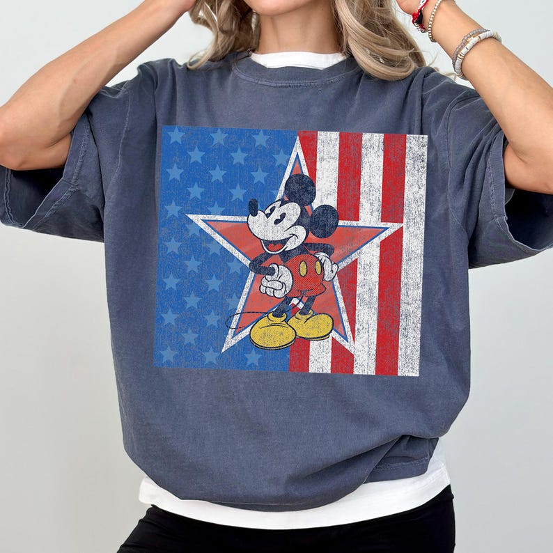Pu&ograve; includere: T-shirt blu con grafica di Mickey Mouse davanti a una stella rossa e al design della bandiera americana. Mickey indossa pantaloncini rossi, scarpe gialle e guanti bianchi. La maglietta ha un aspetto vintage.