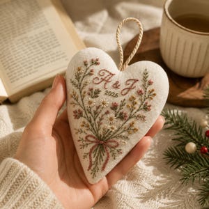 May include: A heart-shaped fabric ornament featuring intricate embroidery of a wildflower bouquet in shades of pink, yellow, and green. The design includes the embroidered letters A & J and the year 2026, with a braided cord loop for hanging.