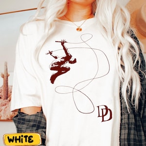 May include: White t-shirt featuring a brown graphic of a figure swinging on a rope, with the letters "DD" in the lower right corner. The shirt has a crew neck and short sleeves. The word "WHITE" is in a yellow rectangle at the bottom.