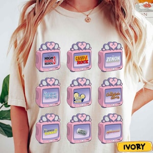May include: Ivory-colored t-shirt featuring a repeating pattern of pink retro televisions. Each TV screen displays a different movie or show title, including "High School Musical 3," "Camp Rock," and "Zenon." The design includes a heart-shaped antenna.