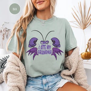 Puede incluir: Una camiseta verde claro con un cangrejo de dibujos animados morado en ella. El cangrejo tiene el texto "Me com&iacute; a mi abuela" escrito debajo.