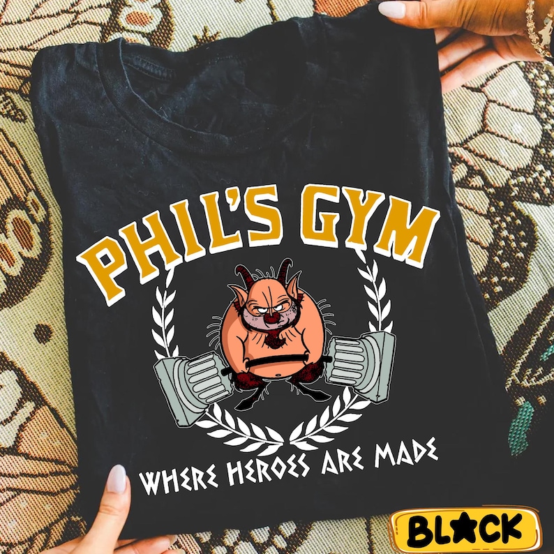 May include: Black t-shirt featuring the text "PHIL'S GYM" in gold and white, with an illustration of a cartoon character lifting weights. The phrase "WHERE HEROES ARE MADE" is below the illustration. A small, yellow "BLACK" tag is in the bottom right corner.