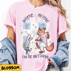 Pode incluir: T-shirt rosa claro com uma ilustra&ccedil;&atilde;o de desenho animado de personagens de Zootopia. O design inclui o texto "WHERE ANYONE CAN BE ANYTHING", pegadas e esbo&ccedil;os. O design &eacute; l&uacute;dico e colorido.