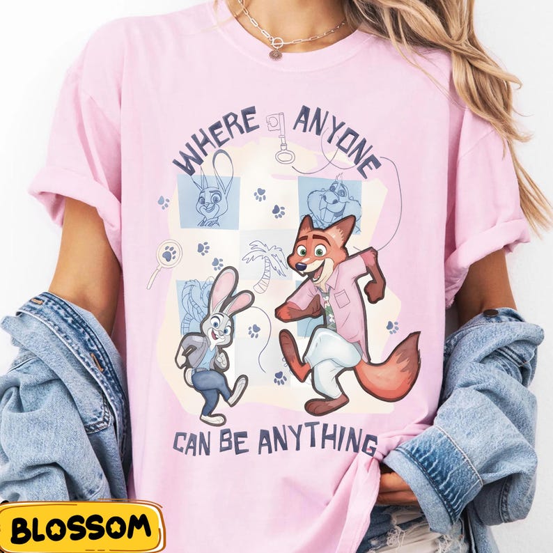 Pode incluir: T-shirt rosa claro com uma ilustra&ccedil;&atilde;o de desenho animado de personagens de Zootopia. O design inclui o texto "WHERE ANYONE CAN BE ANYTHING", pegadas e esbo&ccedil;os. O design &eacute; l&uacute;dico e colorido.