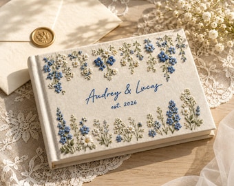 May include: A rectangular cream-colored fabric-covered book featuring embroidered blue and white wildflowers along the top and bottom edges. The center displays the blue embroidered text Audrey & Lucas and est. 2026 in a cursive and print font.