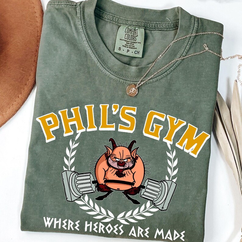 May include: Olive green t-shirt with the text "PHIL'S GYM" in yellow and white, featuring a cartoon character lifting weights. The shirt also has the text "WHERE HEROES ARE MADE". A gold necklace with a pendant is on the shirt.