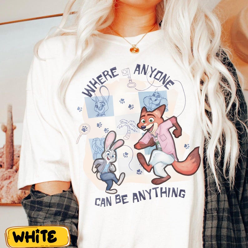 Pode incluir: Camiseta branca com uma ilustra&ccedil;&atilde;o de desenho animado de uma raposa e um coelho de Zootopia, com o texto "WHERE ANYONE CAN BE ANYTHING". O design inclui pegadas e uma chave. A camiseta tem gola redonda e mangas curtas.