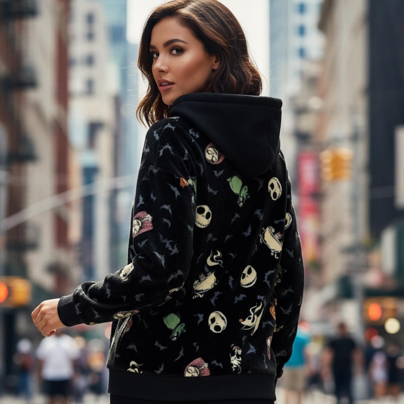 May include: Black hooded sweatshirt featuring a pattern of cartoon characters and bats. The sweatshirt has a black hood and cuffs. The design includes images of a skull, a green creature, and other characters. The background is a blurred city street.