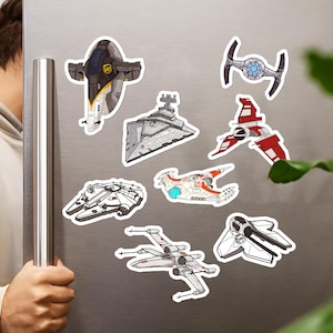 May include: A collection of colorful spaceship stickers, including the Millennium Falcon, X-wing, and TIE fighter, are displayed on a silver refrigerator door. The stickers feature detailed illustrations of various spacecraft.