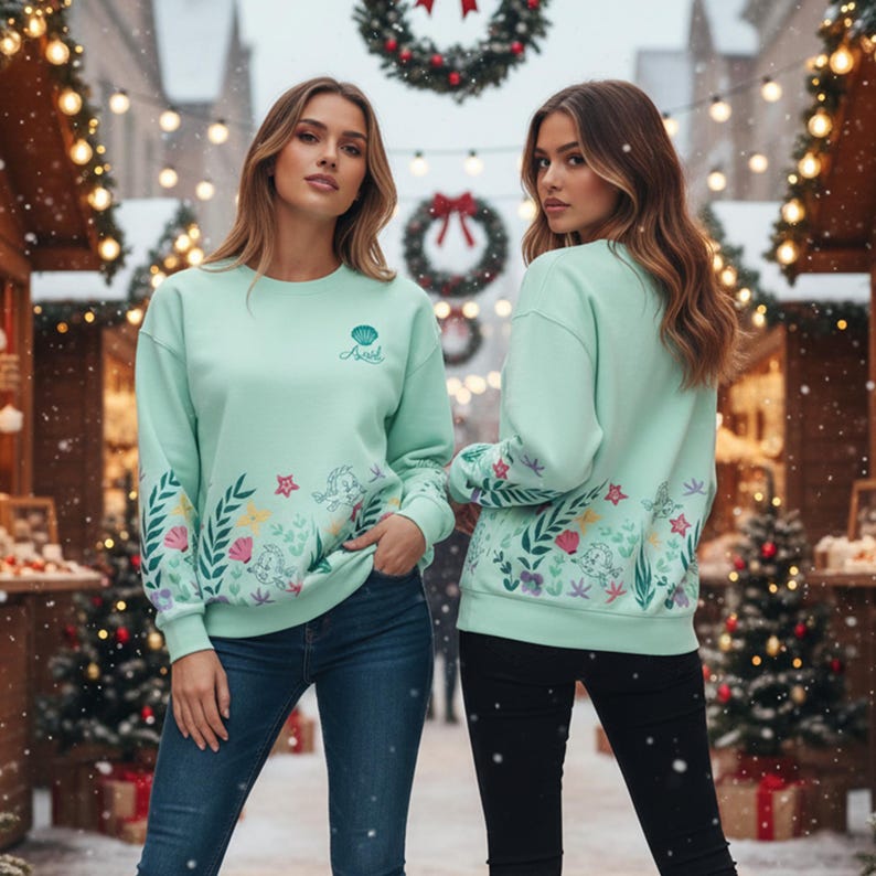 May include: Two mint green sweatshirts with floral and seashell designs. The sweatshirts have the word "Ariel" written on the front. The sleeves and lower portion of the sweatshirts are decorated with colorful flowers and sea elements. The models are wearing blue jeans and black pants.