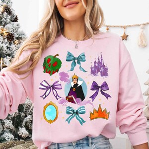 May include: Pink sweatshirt featuring various graphic elements. The design includes a green apple, bows, a castle, a queen, a mirror, and a crown. The graphics are in shades of green, purple, blue, and gold.