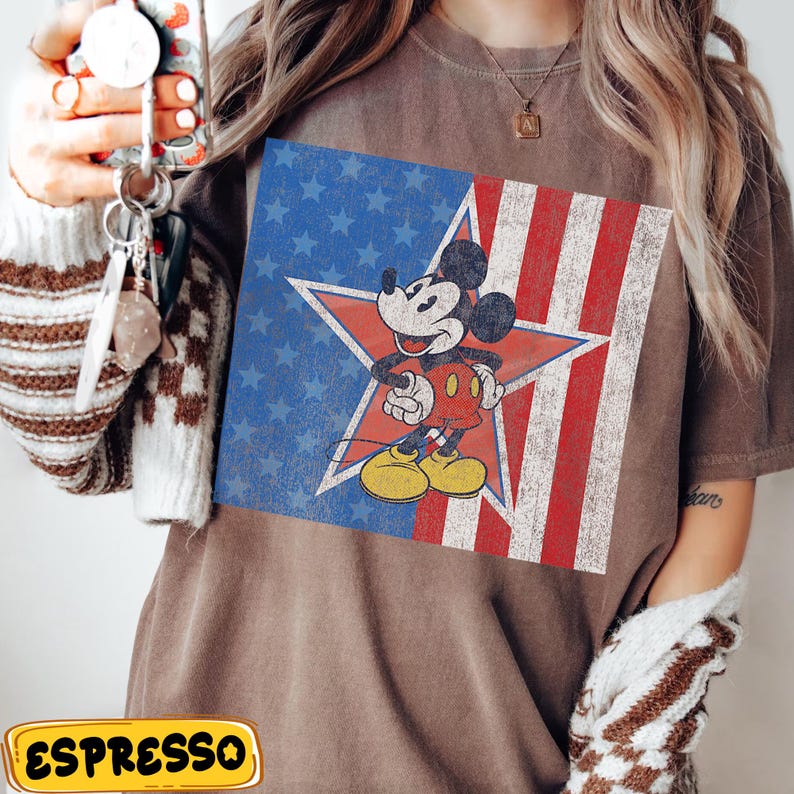 Pu&ograve; includere: T-shirt marrone con una grafica vintage di Mickey Mouse all'interno di un design a stelle e strisce. Il design utilizza rosso, bianco e blu, evocando un tema patriottico. La maglietta ha maniche corte e una vestibilit&agrave; rilassata.