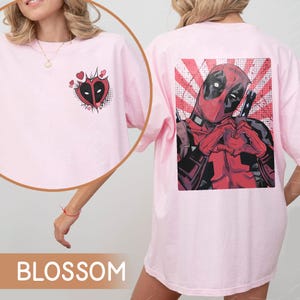 May include: Pink t-shirt with a graphic of Deadpool on the back and a heart with Deadpool's mask on the front. The text "BLOSSOM" is on the bottom of the image.