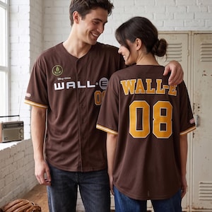 May include: Brown baseball jersey with "WALL-E" and the number "08" in gold lettering. The jersey has white and gold trim on the sleeves and a Disney Pixar logo. The jersey is worn by two people.