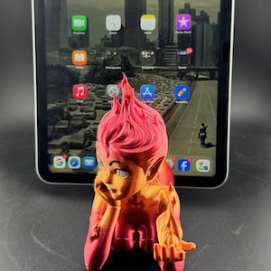 May include: A 3D-printed figurine of a character with pink hair and orange skin, posed in front of a tablet displaying app icons and a cityscape background. The figurine is resting its head on its hand.