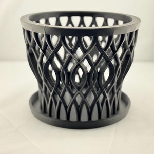 Könnte beinhalten: Schwarzer Plastiktopf mit geometrischem Design. Der Topf hat eine breite Basis und eine schmale Oberseite, wodurch eine Trichterform entsteht.