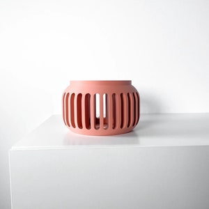May include: A coral-colored ceramic planter with a geometric design. The planter has a cylindrical shape with vertical slits around the sides.