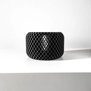 May include: A black, cylindrical, wire mesh planter with a diamond pattern. The planter is empty and sitting on a white surface.
