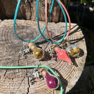 Puede incluir: Cuatro botellas de vidrio con tapones de corcho llenas de purpurina de colores, cada una unida a un cordón con un amuleto. Los amuletos son un dragón, una flor, una estrella y un remolino.