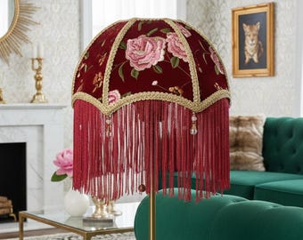 May include: A decorative table lamp with a burgundy shade featuring embroidered pink roses and gold trim. The shade has long red fringe and hangs over a brass base. The lamp is a vintage-inspired home decor item.