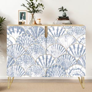 May include: A two-door cabinet with a seashell pattern in blue and white, accented with gold trim. The cabinet has gold hairpin legs and a light wood top. A straw hat hangs on the wall above the cabinet.