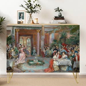 May include: A wooden cabinet with a painted scene of a historical gathering, featuring gold hairpin legs. The artwork depicts a lively outdoor event with numerous figures in period attire. A straw hat and decorative items sit atop the cabinet.