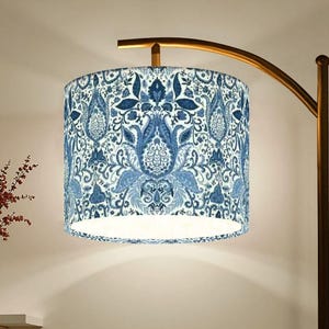 May include: A floor lamp with a curved, gold-toned arm supporting a cylindrical lampshade. The lampshade features a blue and white floral pattern. A small table with a vase of red flowers is in the background.