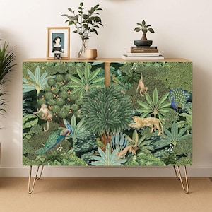 May include: A wooden cabinet with a jungle-themed design featuring monkeys, a leopard, and a peacock. The cabinet has gold hairpin legs and is decorated with lush green foliage. A straw hat hangs above the cabinet.