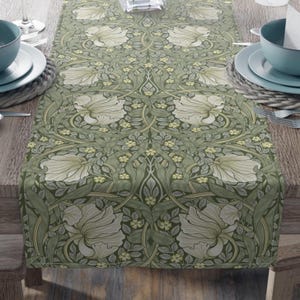 Puede incluir: Un camino de mesa floral verde con un patrón repetido de grandes flores blancas y pequeñas flores amarillas. El camino de mesa está colocado sobre una mesa de madera, con un diseño simétrico.