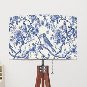May include: A white lampshade with a blue and white floral pattern featuring birds. The lampshade is on a wooden tripod stand.